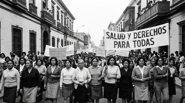 fotografía histórica en blanco y negro de una marcha de mujeres en el Perú con pancartas y diversidad de edades, enfoque en rostros y cartelería